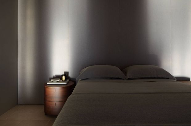 A dark, minimalist bedroom with a low-profile bed dressed in charcoal bedding, set against full-height dark grey wall panels. A small rounded timber bedside table with a simple lamp and dish sits to the left. The room has a restrained, moody palette and minimal furnishings.