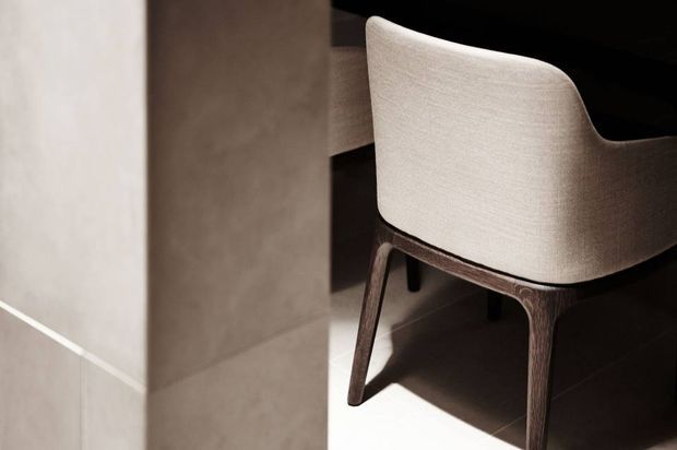 Close-up interior detail of a sculptural upholstered dining chair with a rounded cream fabric shell and dark timber legs, positioned beside a dark table or built-in surface. Neutral stone or plaster wall surfaces and tiled flooring are visible, with strong directional light creating deep shadows. The image emphasises refined material contrast and furniture form rather than a complete room.