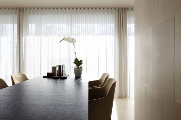 A refined dining room with a large dark dining table and upholstered dining chairs positioned in front of full-height sheer curtains. The room features abundant diffused daylight, pale floor and wall finishes, and a simple orchid centerpiece. Large-format wall panels and clean-lined furnishings create an understated, contemporary setting.