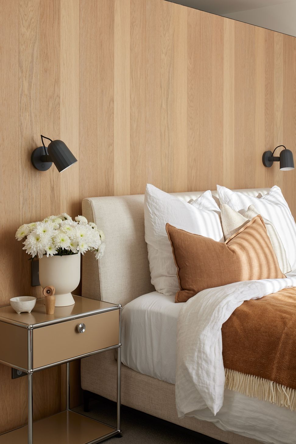 The main bedroom is designed as a peaceful retreat, featuring a built-in oak headboard, soft white and tan bedding, and black wall-mounted reading lights.