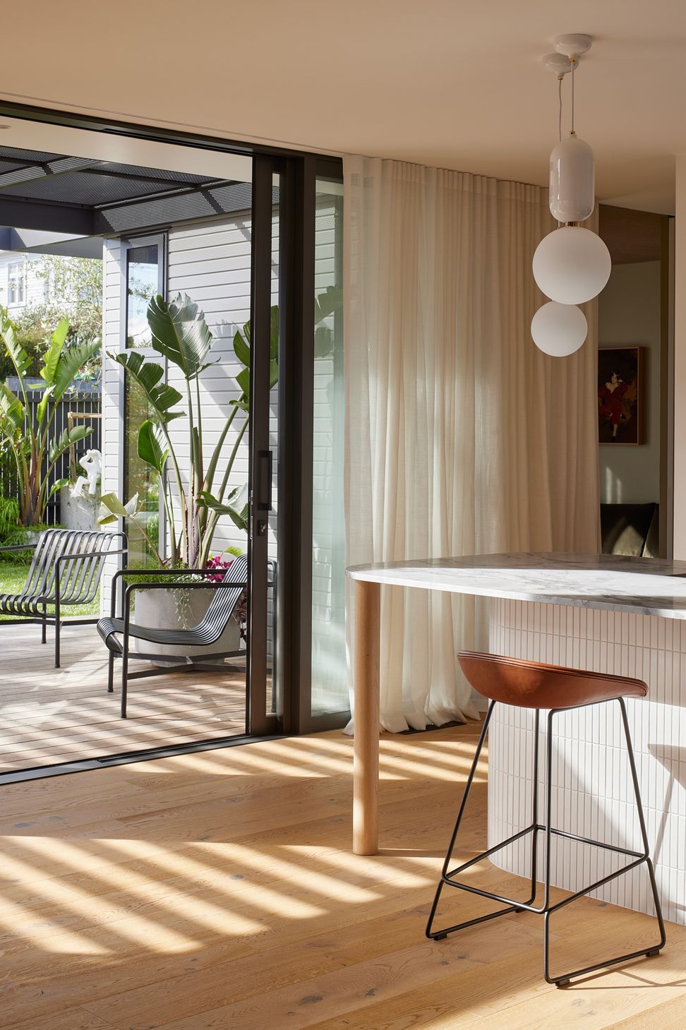 The kitchen island showcases its rounded oak detailing beneath feature pendant lights. Beyond, the deck with Hay outdoor lounge furniture extends the living spaces into the garden.