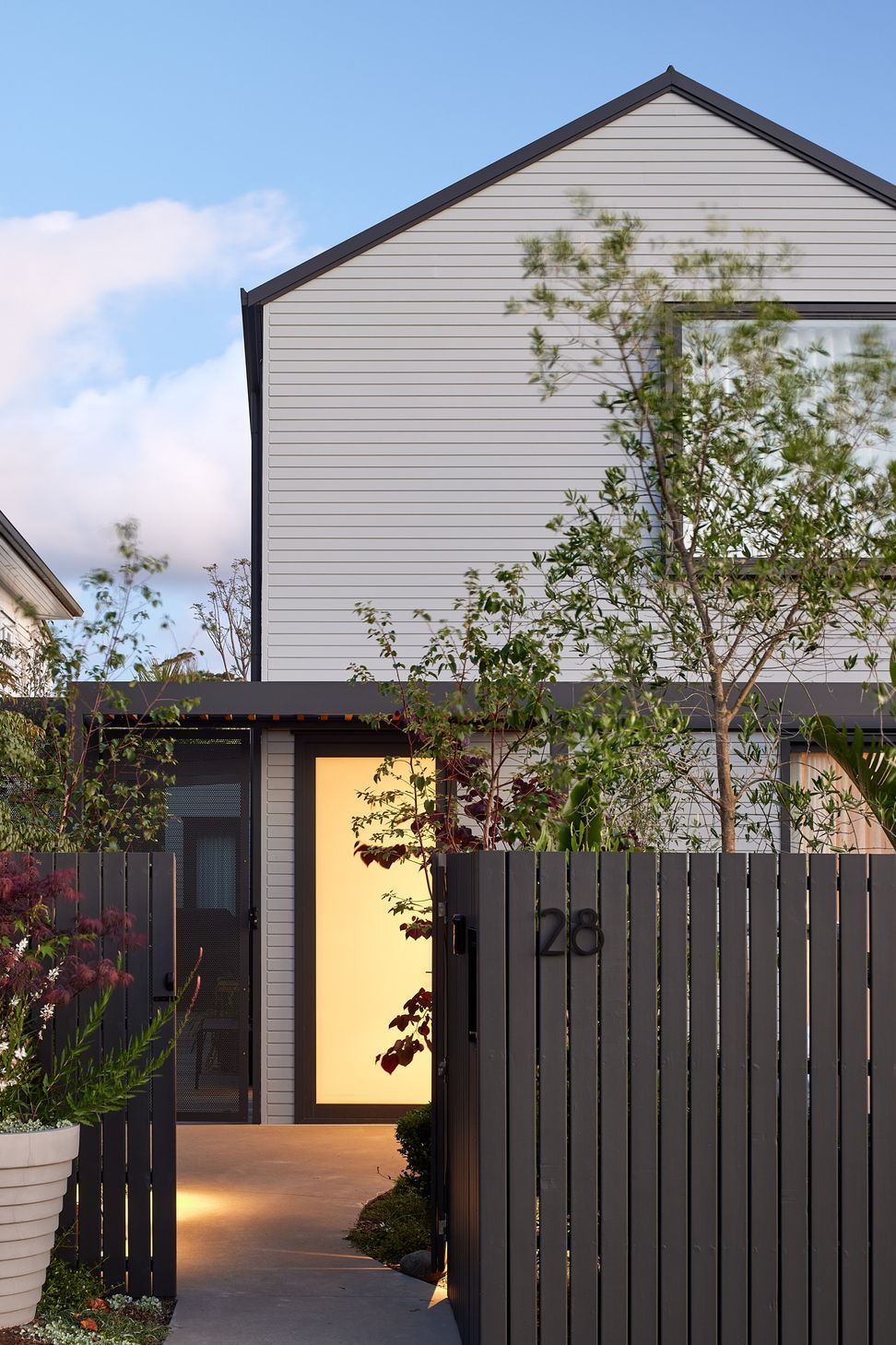 At dusk, the approach to the home is both welcoming and understated. A softly illuminated front door with opaque glazing is framed by mature planting and a dark grey timber fence.