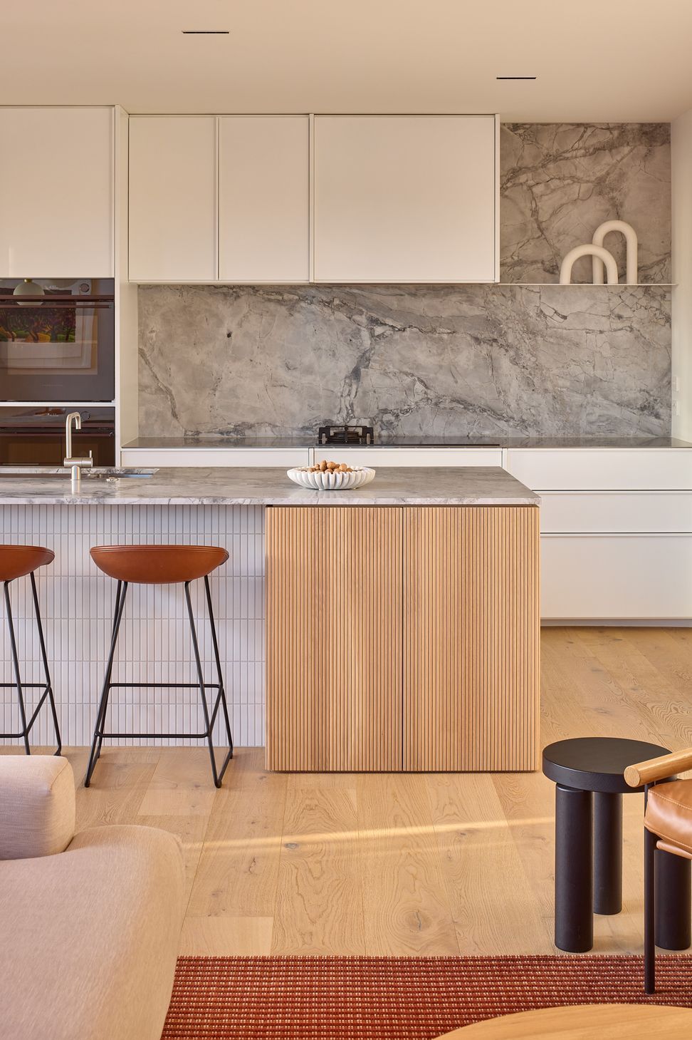 Viewed from the living room, the kitchen is defined by its sculptural island, bespoke rear cabinetry and restrained material palette. The composition balances functionality with refined detailing.