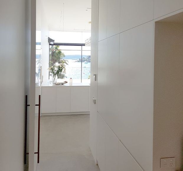 Bright minimalist interior viewed through an open doorway, with full-height white built-in cabinetry, pale flooring, and a large glazed opening overlooking water and a coastal landscape. A narrow glimpse of a dining or living area is visible beyond the cabinetry.