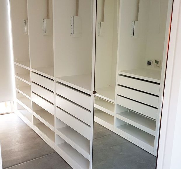 Bright walk-in wardrobe with extensive floor-to-ceiling white built-in shelving, hanging compartments, and handleless drawers. A full-height mirror reflects the cabinetry, while the polished concrete floor and minimal detailing create a clean, contemporary fit-out.