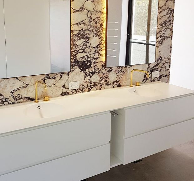 Contemporary double vanity bathroom with a long white integrated basin and two white drawer vanities. The wall behind the vanity is clad in dramatic veined natural stone, complemented by slim gold-toned wall-mounted tapware, mirrored cabinetry, and warm concealed lighting.