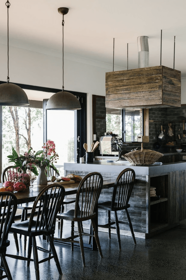 A rustic, open-plan kitchen and dining area with a large island clad in weathered timber, dark cabinetry, and a substantial timber rangehood surround. Black industrial pendant lights hang above a dark dining table with spindle-back chairs. The kitchen includes dark tiled walls, a polished concrete floor, stainless-steel ventilation ducting, and large windows overlooking trees. Natural floral arrangements and timber kitchen accessories add warmth.