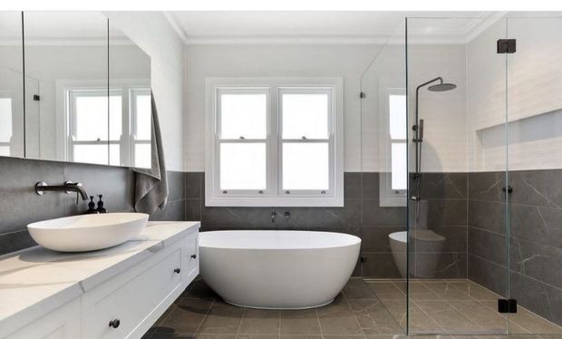 Bright contemporary bathroom featuring a freestanding white oval bathtub beneath a double window, a white vessel sink on a built-in vanity, and a frameless glass walk-in shower. The room has two-tone walls with white upper surfaces and dark grey tiled lower walls, matching dark grey floor tiles, mirrored cabinetry, and polished chrome fixtures.
