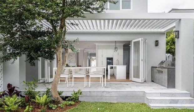 A bright contemporary outdoor entertaining area adjoining a white rendered house, with a covered patio, dining table and chairs, open bi-fold doors, a built-in outdoor kitchen with grill, tiled steps, and landscaped planting. The interior kitchen is visible through the open doors.