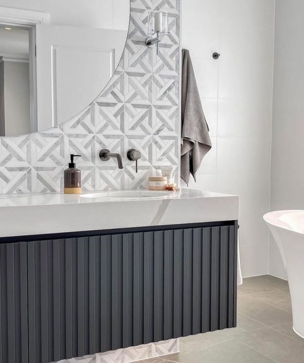 A bright contemporary bathroom featuring a wall-mounted dark vertical-groove vanity with a white stone benchtop, brushed dark tapware, and a geometric grey-and-white tiled splashback that continues around the mirror. A freestanding white bathtub is partially visible at right, with large light-grey wall and floor tiles and a neutral towel adding texture.