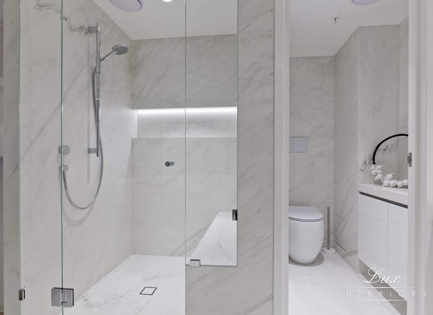 Bright contemporary bathroom featuring a frameless glass walk-in shower, wall-mounted rain shower and hand shower, continuous pale marble-look wall and floor surfaces, a large mirrored panel with concealed linear lighting, and a separate toilet and floating vanity zone.