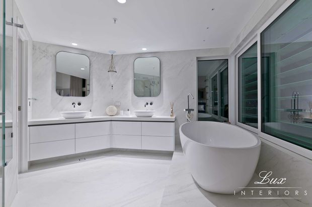 A bright, spacious contemporary bathroom with a double floating vanity, two rounded rectangular mirrors, wall-mounted basins, a freestanding oval bathtub, and large glazed windows. The room is finished in a predominantly white and pale grey palette with marble-look wall and floor surfaces, streamlined white cabinetry, chrome tapware, and recessed ceiling lighting.