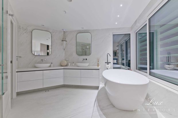 Bright, spacious contemporary bathroom with a freestanding white bathtub positioned beside large windows, a double floating vanity with two vessel basins, rounded rectangular mirrors, pale marble-look wall and floor finishes, and frameless glass shower glazing. The room has recessed ceiling lights and a clean white-grey palette.