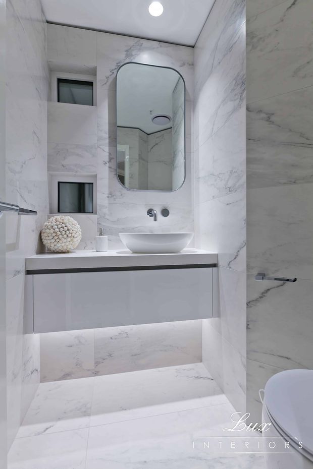 A compact, bright bathroom clad in white marble-look tiles with grey veining. It features a floating white vanity with an integrated countertop, vessel basin, wall-mounted chrome tapware, a rounded rectangular mirror, recessed shelving, and concealed under-vanity lighting. A partial toilet is visible at the right edge.