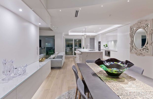 Bright, open-plan contemporary interior combining a dining area, kitchen and living space. The room features white built-in cabinetry, pale timber flooring, a stone kitchen island with bar stools, a grey dining table with upholstered chairs, recessed lighting and a curved ceiling detail. An ornate framed mirror adds a decorative traditional accent.