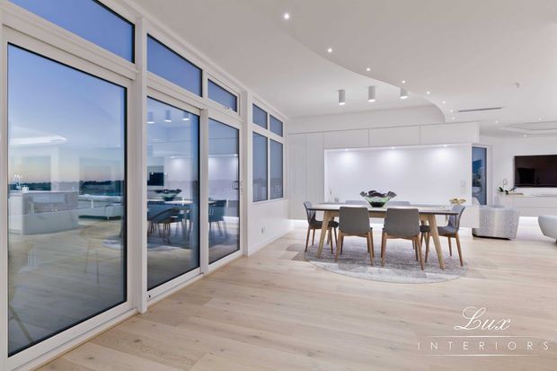 Bright, spacious contemporary open-plan dining and kitchen interior with pale timber flooring, extensive floor-to-ceiling glazing, white built-in cabinetry, a large dining table, and recessed and pendant lighting. The glazing opens toward a bright outdoor view and reflects additional dining furniture. A living area with a television is visible in the background.