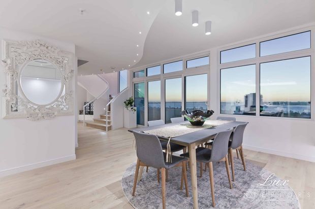 Bright, spacious open-plan dining area with a contemporary dining table and upholstered chairs, pale timber flooring, large framed windows overlooking a city and water view, a sweeping curved ceiling, sculptural pendant lights, and a staircase leading to an upper level. An ornate wall mirror adds a decorative traditional element.