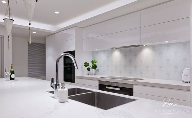 Bright contemporary white kitchen with extensive glossy handleless cabinetry, a large white stone island with integrated double stainless-steel sink, built-in black appliances, and a subtle patterned light backsplash. Recessed ceiling lighting and decorative pendant lights create a clean, refined finish.