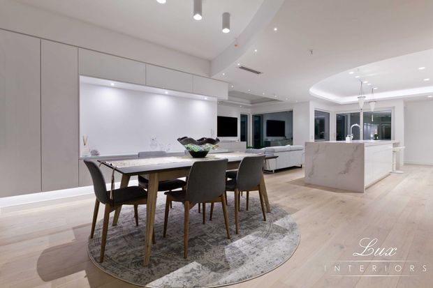 Open-plan contemporary kitchen, dining and living area with pale timber flooring, sleek light-grey full-height cabinetry, a marble-look island bench, and a timber dining table with upholstered chairs. Recessed and pendant lighting, large glazing, and neutral finishes create a bright, refined interior.