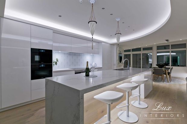 A bright, contemporary open-plan kitchen with extensive glossy white cabinetry, integrated black appliances, a large pale stone island with waterfall ends, and white sculptural bar stools. The room features light timber flooring, a softly curved recessed ceiling with concealed cove lighting, pendant lights, and glazed doors opening toward a dining area.