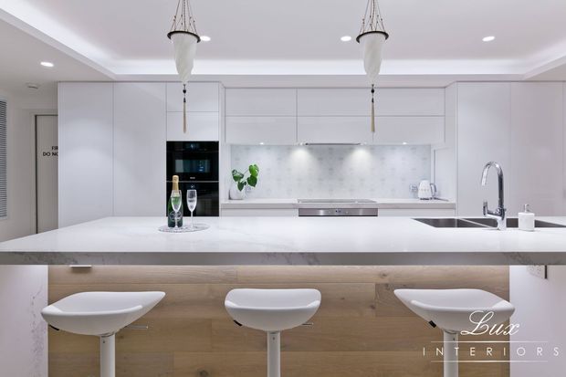 Bright contemporary white kitchen with extensive handleless cabinetry, a pale marble-look splashback, a large white island with integrated sink and curved chrome tap, and a timber-faced breakfast bar with three white stools. Decorative pendant lights and under-cabinet lighting add refined detailing.