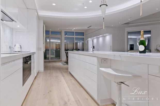 Bright, spacious contemporary white kitchen with extensive built-in cabinetry, a large central island, waterfall-style pale stone benchtops, integrated appliances, and a curved bulkhead ceiling. Pale timber flooring, glass doors, recessed lighting, and white bar seating create a clean, refined interior. A champagne bottle sits on the island, and the image includes a Lux Interiors watermark.