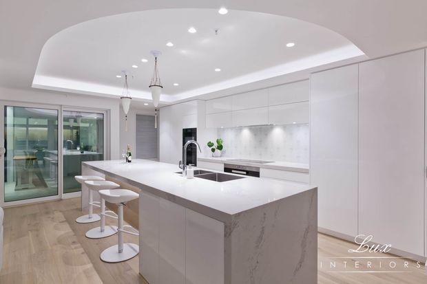 Bright, spacious contemporary white kitchen with a large waterfall-edge island, integrated cabinetry, pale stone surfaces, under-cabinet lighting, pendant lights, and four white bar stools. Glazed sliding doors open to an adjoining outdoor or living area, while light timber flooring adds warmth.