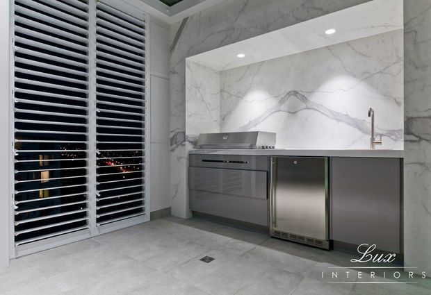 Contemporary compact kitchen with dramatic white marble-look stone wrapping the walls and splashback, integrated stainless-steel appliances, dark grey cabinetry, recessed ceiling lighting, and full-height white louvred shutters overlooking city lights.