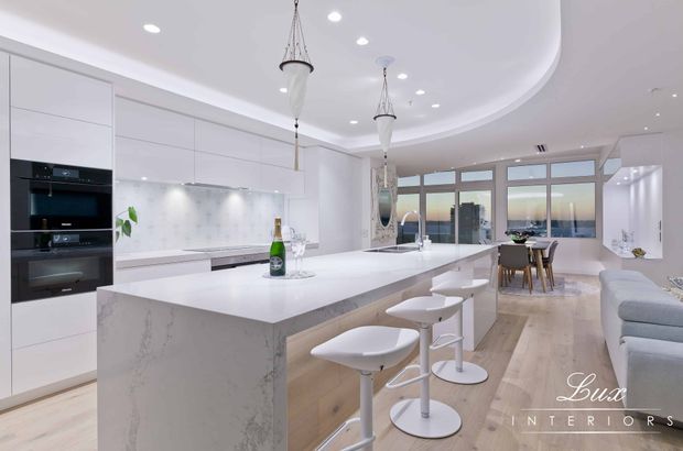Bright, spacious open-plan kitchen and living area with a large white stone island, integrated white cabinetry, black built-in appliances, pendant lights, timber-look flooring, bar stools, dining area and floor-to-ceiling glazing. The interior has a clean, contemporary coastal feel with a curved ceiling detail and water views or bright exterior beyond the windows.