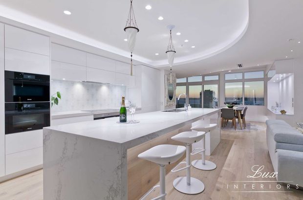 Bright, spacious contemporary open-plan kitchen and living area with a large white waterfall island, integrated appliances, white cabinetry, pale timber flooring, pendant lights, and bar stools. Full-height glazing opens to an outdoor view, while a dining area and sofa are visible beyond the kitchen.