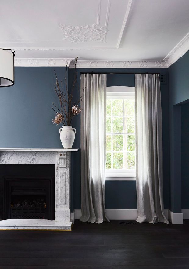 A refined traditional living room with deep blue walls, ornate white ceiling cornices and plaster ceiling medallion, a classic marble fireplace, dark timber flooring, and tall windows dressed with full-length grey curtains. The room combines heritage architectural detailing with a restrained, sophisticated palette.