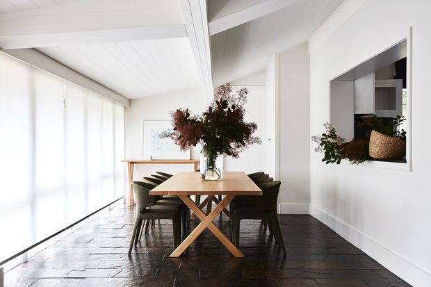 Bright dining area with a rectangular timber dining table and dark dining chairs, set against white walls and a white paneled ceiling. Large translucent floor-to-ceiling glazing provides soft natural light, while dark timber flooring and exposed white ceiling beams add contrast. A large floral arrangement sits at the centre of the table, and a recessed wall opening reveals part of an adjacent kitchen or display area.