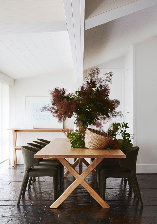 Bright dining room with a substantial natural timber trestle table surrounded by dark upholstered dining chairs. The room has white painted paneled walls and a white vaulted, exposed-beam ceiling, dark timber flooring, and a slim console table against the wall. A large leafy floral arrangement in a woven basket forms the centrepiece, creating an organic and refined atmosphere.