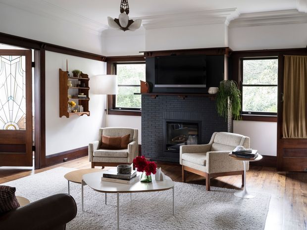 A refined living room with dark timber-framed windows and door, white walls, ornate ceiling cornices, and a central dark brick fireplace with a mounted television. Two upholstered lounge chairs, a round coffee table, a large light rug, shelving, indoor plants, and warm timber flooring create a comfortable, traditional-meets-contemporary interior.