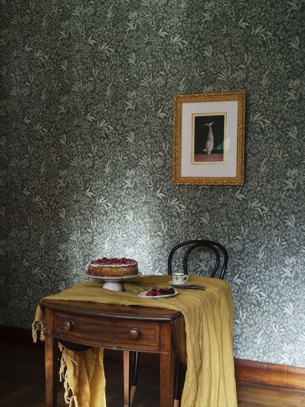 A moody, traditionally styled dining vignette featuring a dark botanical patterned wallpaper, an antique wooden console or dining table draped with a mustard textile, a black bentwood chair, pastries and tableware, and a small ornate gold-framed artwork. Dark timber flooring and timber skirting reinforce the warm, historic character.