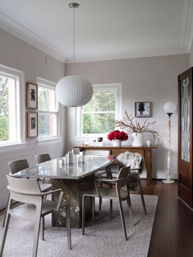Bright dining room with a rectangular marble dining table, contemporary upholstered and molded chairs, a large spherical pendant light, white-painted brick walls, dark timber flooring, and tall windows overlooking greenery. A timber console, decorative branches, artwork, and a glass-fronted cabinet add character to the refined interior.