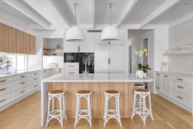 Bright, spacious contemporary kitchen with a large central island clad in vertical light timber battens, white cabinetry, integrated appliances, timber flooring, and four white bar stools. The room features a coffered or boxed-beam ceiling, twin oversized pendant lights, open shelving, extensive built-in storage, and a neutral white-and-light-wood palette.