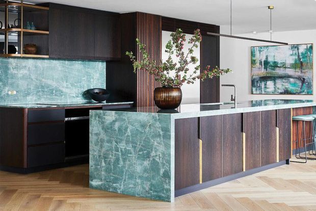A sophisticated contemporary kitchen featuring dark timber cabinetry, a large green-veined stone island and matching splashback, integrated shelving, vertical timber screening, brass-toned pendant lighting, and warm herringbone timber flooring. A sculptural vase and artwork add refined decorative accents.