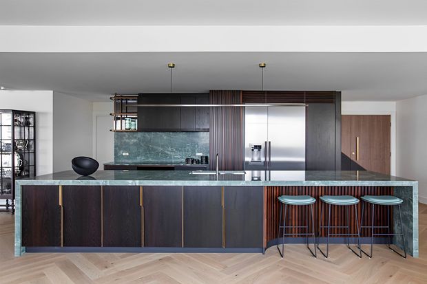 Contemporary open-plan kitchen dominated by a long dark timber island with a green-toned stone benchtop and integrated seating for three. The kitchen includes dark cabinetry, stainless-steel appliances, a textured green stone splashback, open shelving, pendant lighting, and warm herringbone timber flooring. A dining or living area is partially visible beyond the kitchen.