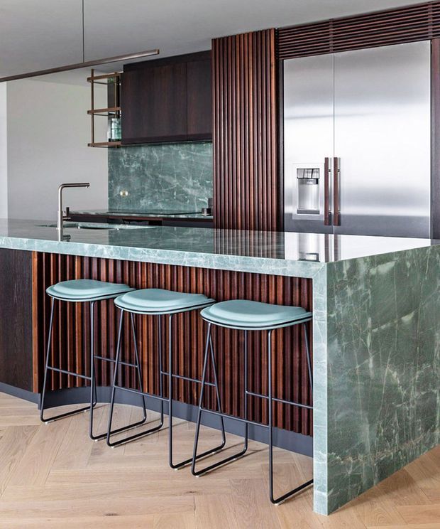 Contemporary kitchen featuring a large green-veined stone island with integrated sink and waterfall end panel, dark timber vertical slatted cabinetry and island cladding, stainless steel refrigerator, and three slim bar stools. Light herringbone timber flooring and a pale ceiling create contrast with the rich cabinetry and stone surfaces.
