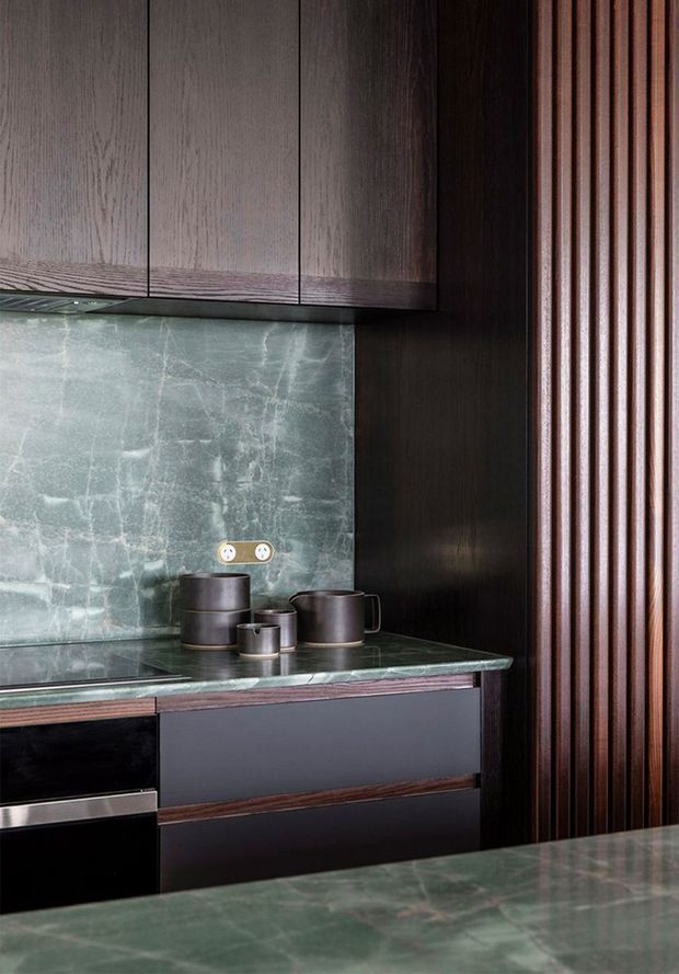 Close-up view of a refined dark-toned kitchen featuring deep timber cabinetry, a green veined stone splashback and benchtop, integrated dark cabinetry, and a vertical timber slat wall detail. Ceramic vessels sit on the counter, reinforcing the moody, luxurious material palette.