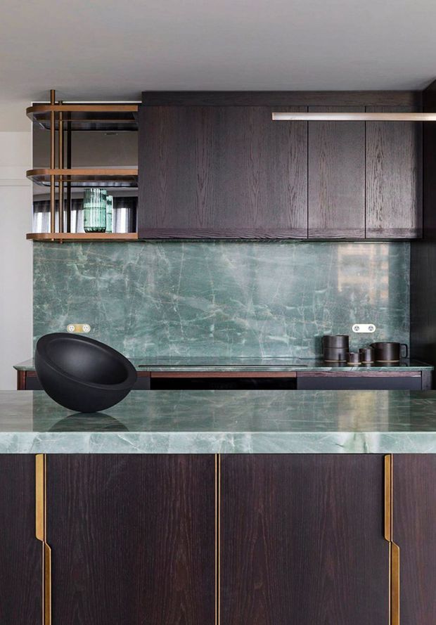Close-up view of a sophisticated dark timber kitchen featuring richly grained cabinetry, a green-grey veined stone splashback and island benchtop, integrated brass detailing, open timber-and-metal shelving, and a black sculptural bowl. The composition is refined and dramatic with a moody, luxe material palette.