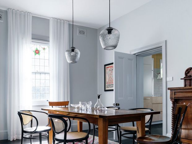 A bright, refined dining room with a dark timber rectangular dining table, woven-seat black-framed chairs, sheer white curtains, a tall leaded-glass window, pale grey walls, and two spherical pendant lights. An open doorway reveals a tiled adjacent room, while an antique dark timber cabinet adds a traditional accent.