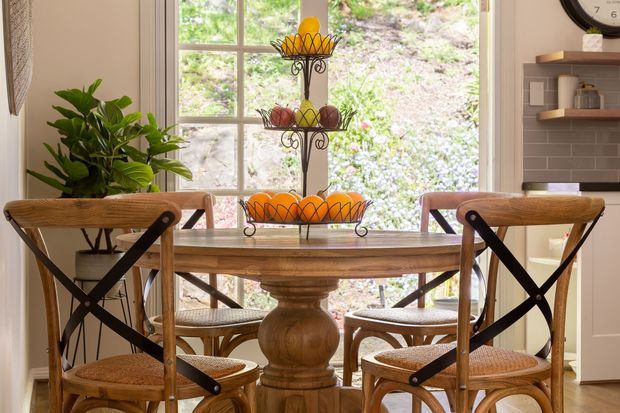 A warm, traditionally styled dining area with a round wooden dining table and matching cross-back chairs with woven seats. A tiered black metal fruit stand forms a central table display, while large multi-pane windows bring in abundant natural light and overlook greenery. A leafy indoor plant, built-in shelving, and a wall clock add detail to the surrounding space.