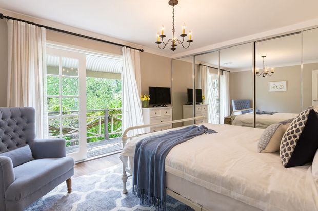 A bright, elegantly styled bedroom with a white upholstered bed, layered neutral bedding, a blue throw, tufted armchair, mirrored built-in wardrobes, white chest of drawers, and a large glazed door opening to a leafy balcony or outdoor area. A traditional chandelier and classic furniture details add refined character.