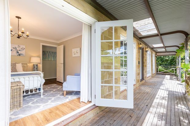 Bright heritage-style home exterior showing a covered timber verandah and deck with a white French door opening into a softly furnished bedroom. The verandah has timber posts, decorative brackets, a corrugated metal ceiling, extensive glazing, and lush greenery beyond. The bedroom includes a white metal bed, pale upholstered chair, patterned rug, timber flooring, neutral walls, and classic decorative details.