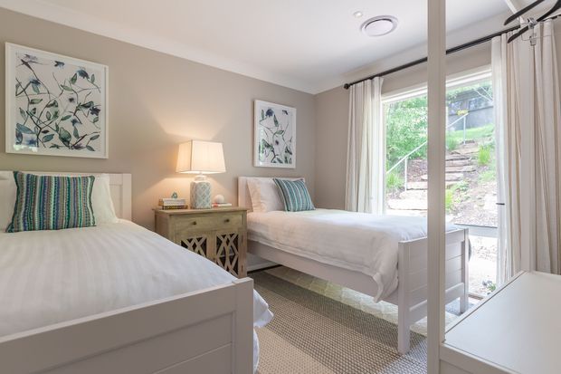 Bright, neatly styled twin bedroom with two white single beds, striped blue-green cushions, a central bedside table and lamps, botanical artwork, neutral taupe walls, and a large glazed opening with light curtains overlooking greenery. The room has a clean, calm and coastal-inspired feel.