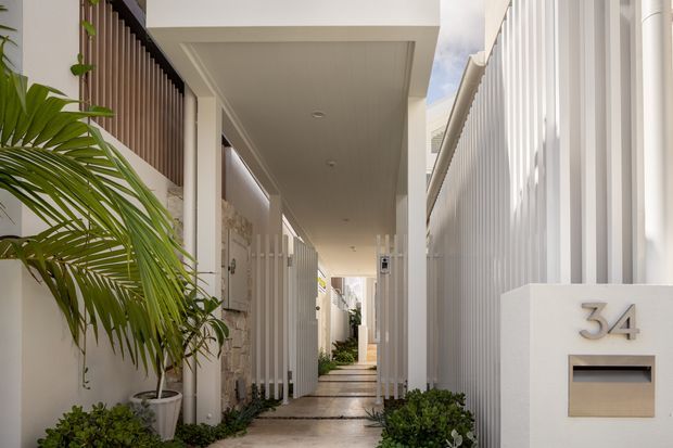 A bright, narrow covered pedestrian entry passage in a contemporary residential development. White rendered walls, vertical privacy screens and slatted fencing line the walkway, with recessed ceiling lights, tropical planting and a house number mailbox creating a clean, refined entrance sequence.