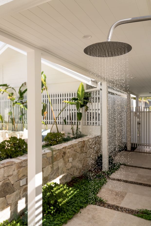 A bright covered outdoor shower area with a large round rainfall showerhead, white-lined ceiling, white structural posts, stone retaining walls, lush planting, and a paved path with pebble drainage strips. The space has a resort-like garden courtyard atmosphere and appears connected to an exterior entry or poolside area.