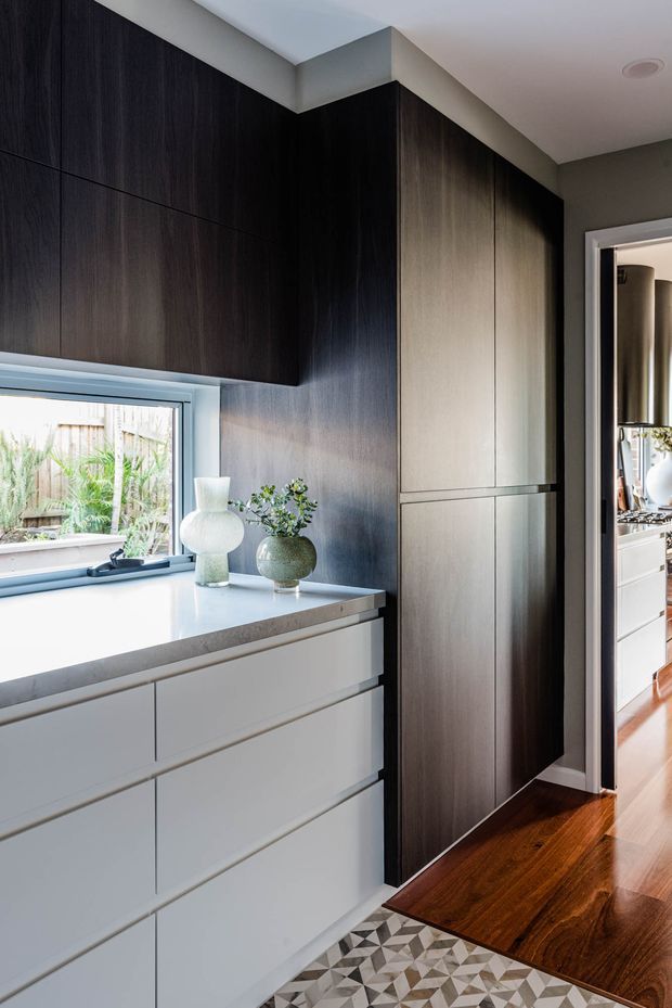 A refined built-in storage and cabinetry area with dark timber-look full-height joinery, pale drawer cabinetry, and a light stone benchtop beneath a window. Warm timber flooring and patterned tile flooring meet at the threshold to an adjoining room visible through the doorway.